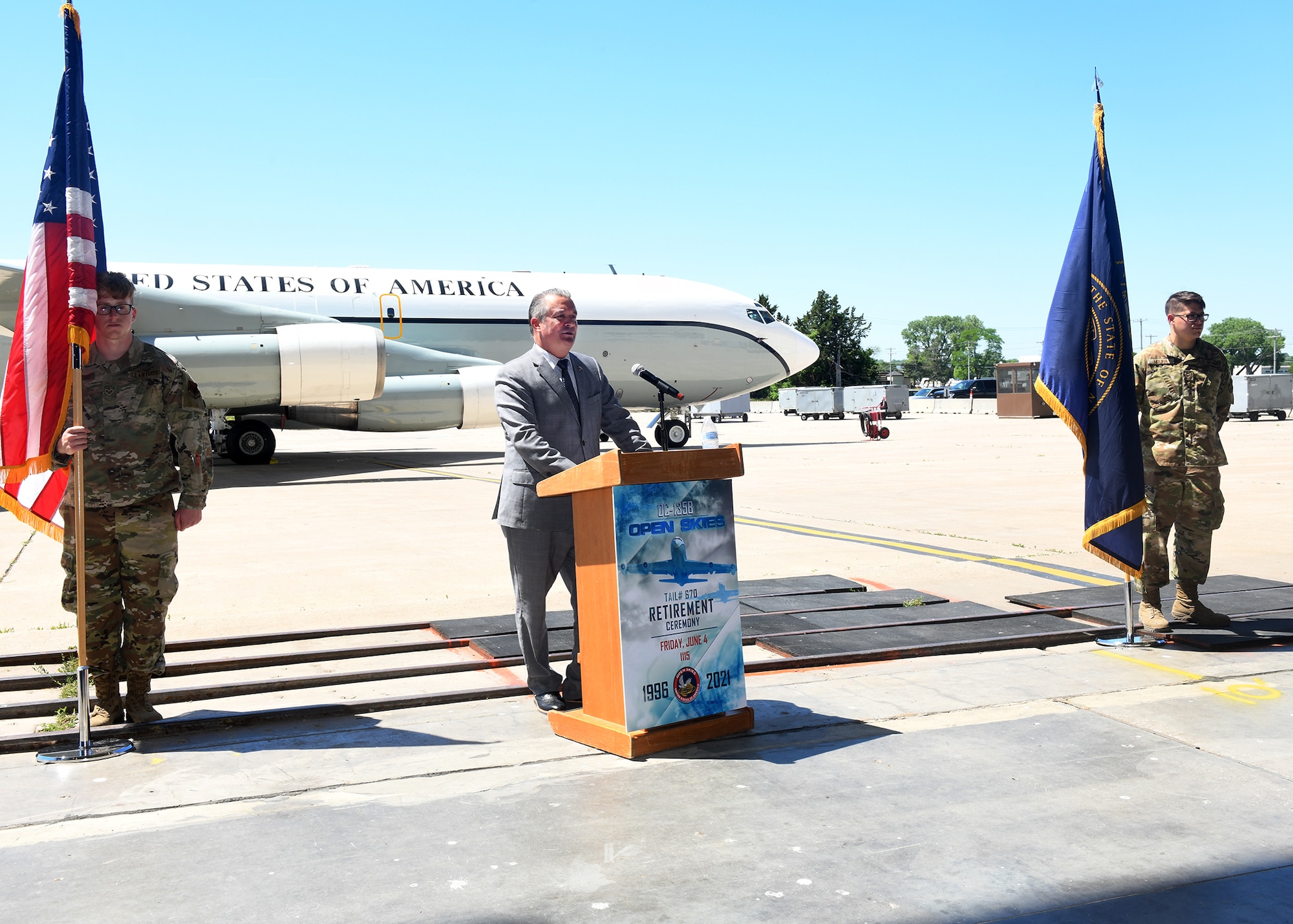 Man gives speech at a podium outside. An Airmen holding a flag stands on each side of the podium and the retiring OC-135 aircraft is in the background.