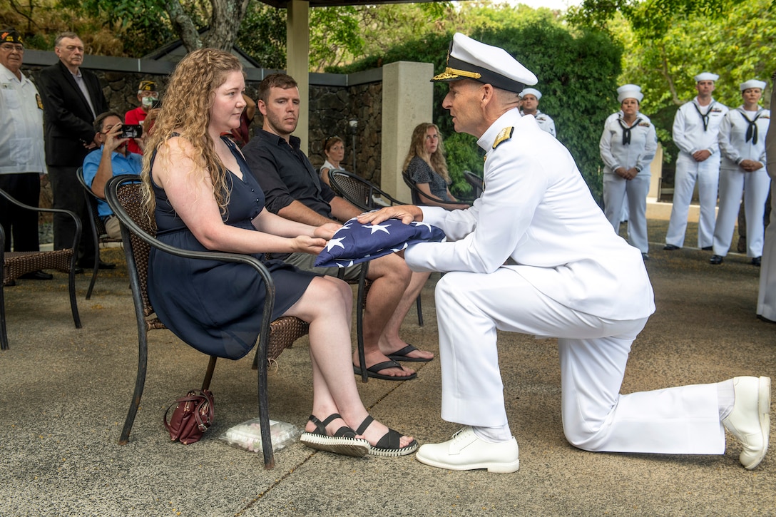 A kneeling sailor presents a folded U.S. flag to a civilian at an outdoor ceremony.