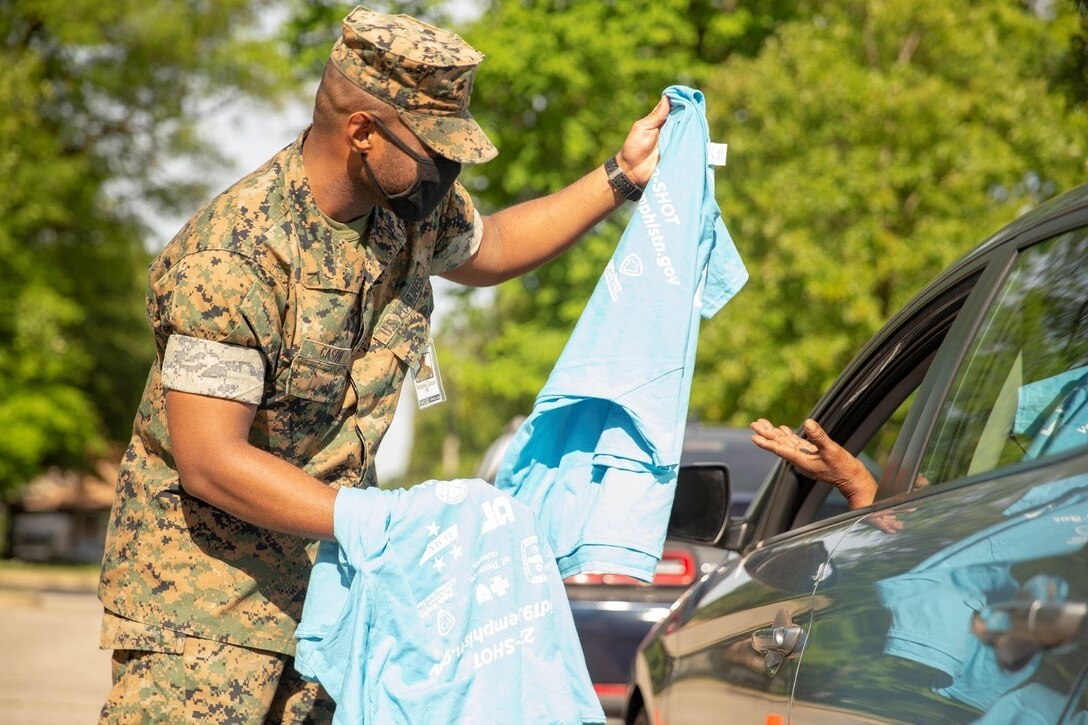 A Marine hands a T-shirt to a person sitting in an automobile.