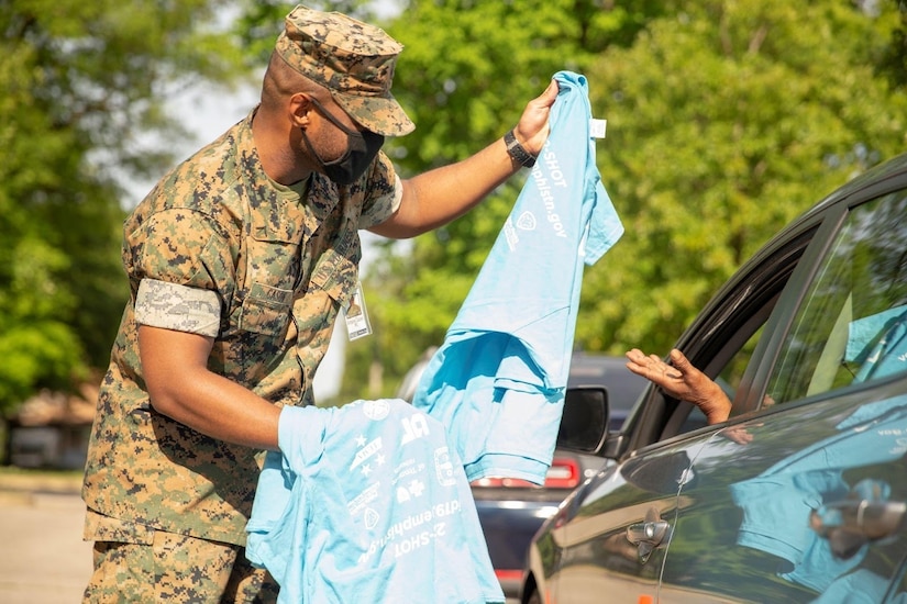 A Marine hands a T-shirt to a person sitting in an automobile.