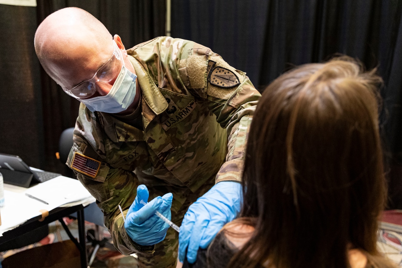 A soldier wears rubber gloves and a mask. He uses a syringe to administer a vaccine into a woman's arm.