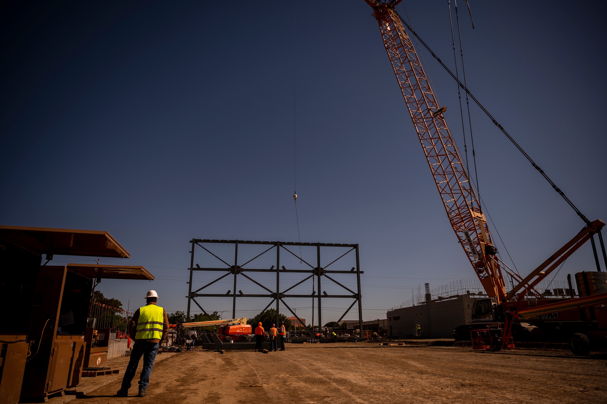 A man is standing in a construction site while a massive crane is being operated and other construction crewmen work.