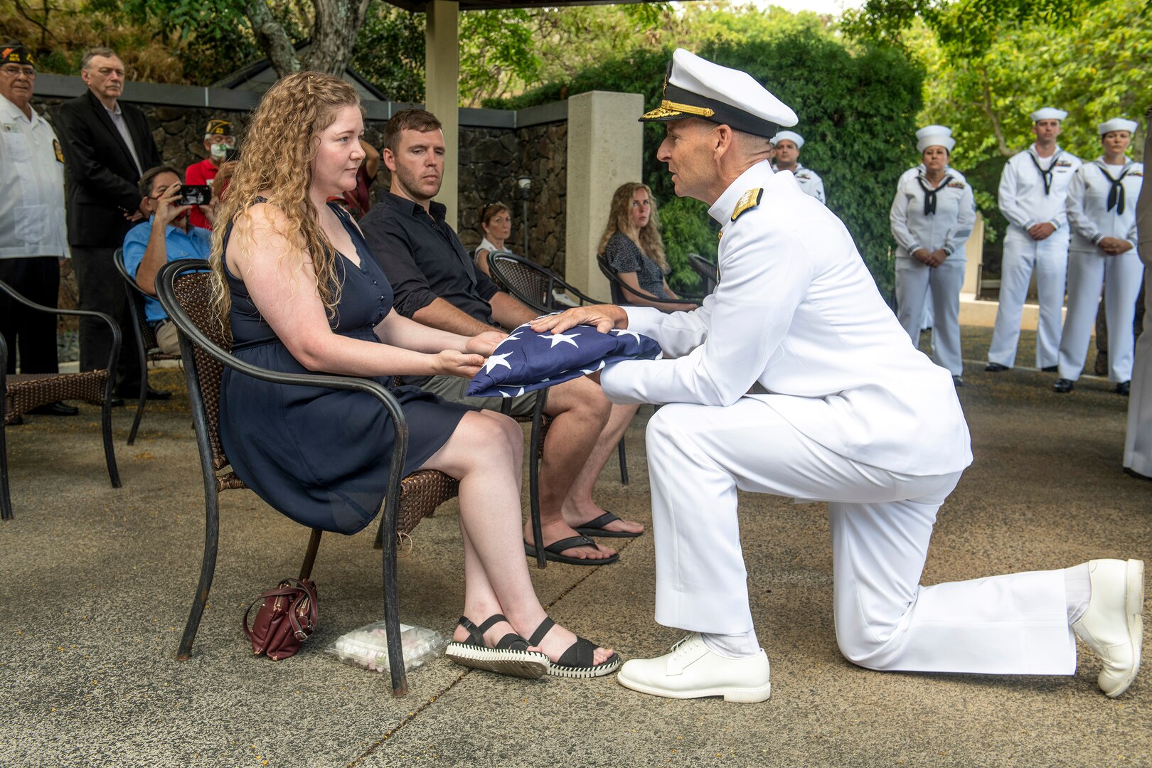 A kneeling sailor presents a folded U.S. flag to a civilian at an outdoor ceremony.