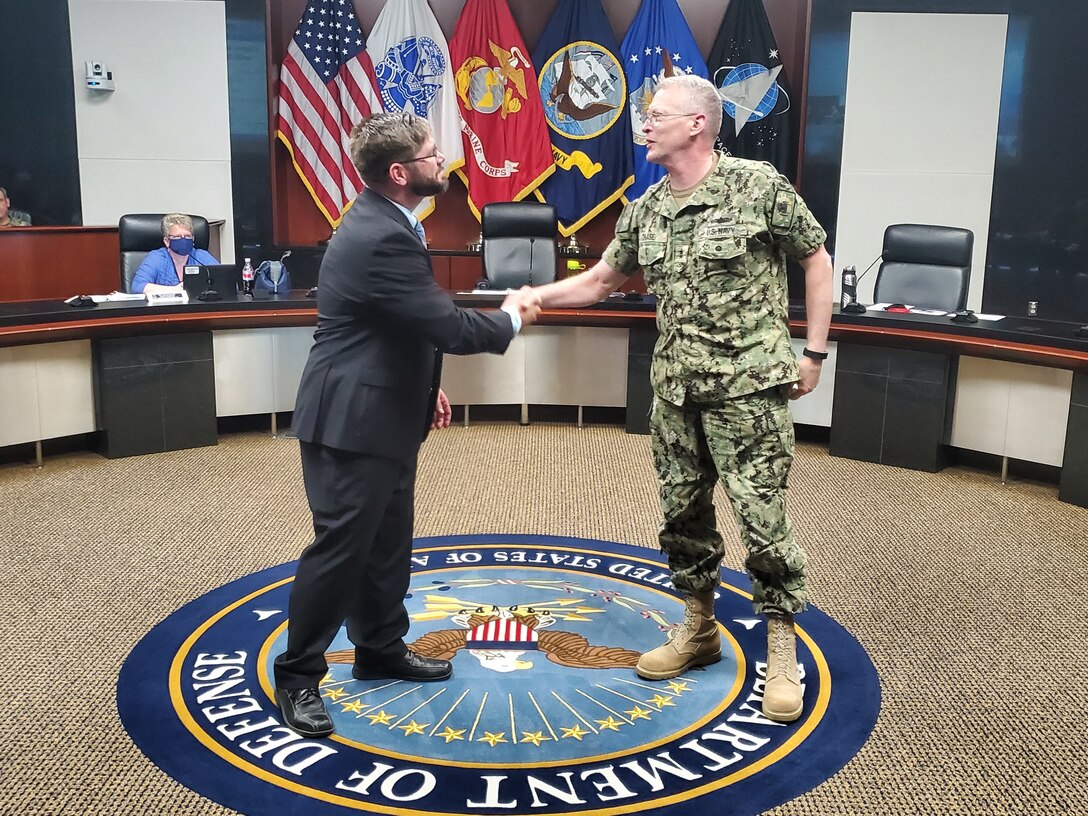 Colin Williams, standing and shaking hands, receives a coin from Navy Rear Admiral Grafton Chase