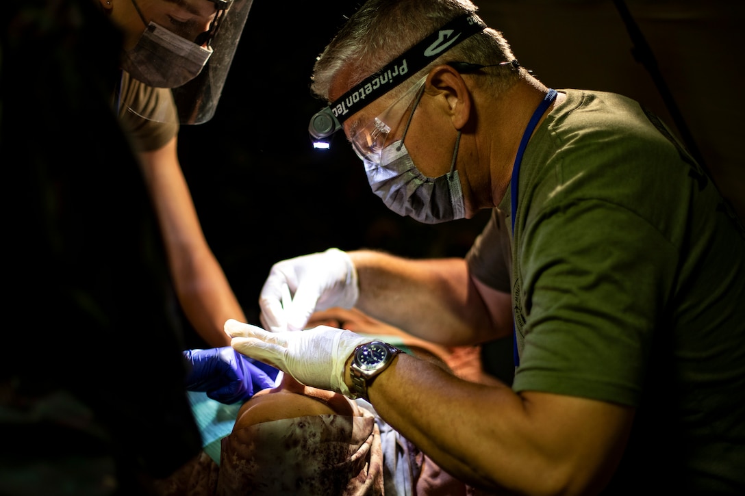 An Air Force dentist works on the mouth of a patient lying supine as a service member stands on the patient's other side.
