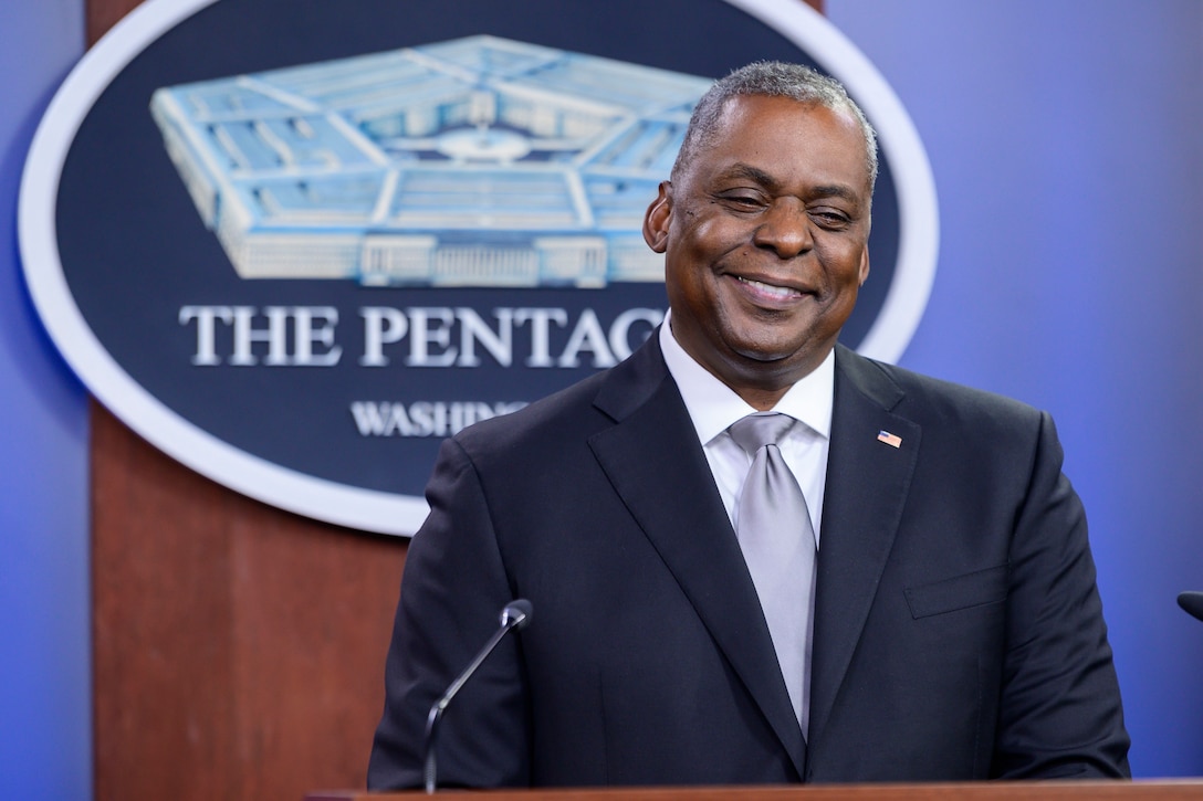 Secretary of Defense Lloyd J. Austin III smiles while standing in front of a microphone.