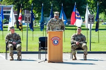 two men sitting wearing army uniforms, one man in uniform standing at a podium.