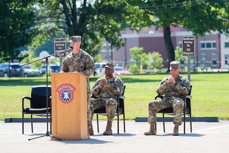 two men sitting wearing army uniforms, one man in uniform standing at a podium.