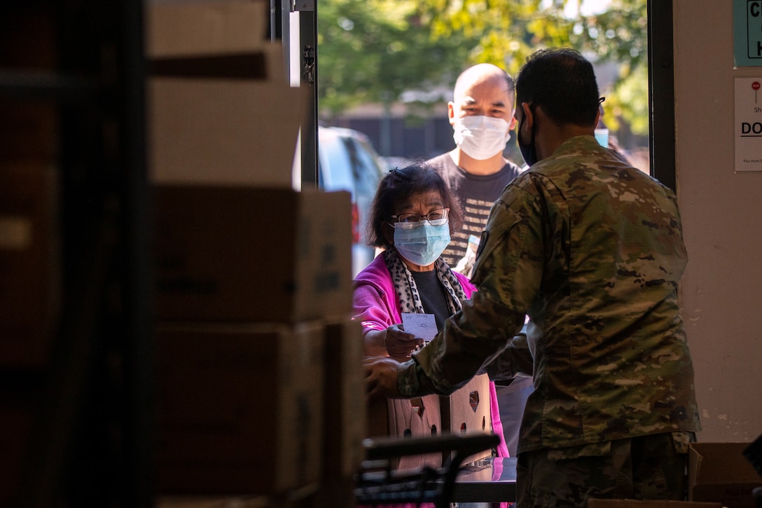 From behind, a  soldier is seen handing a box to a woman wearing a face mask.