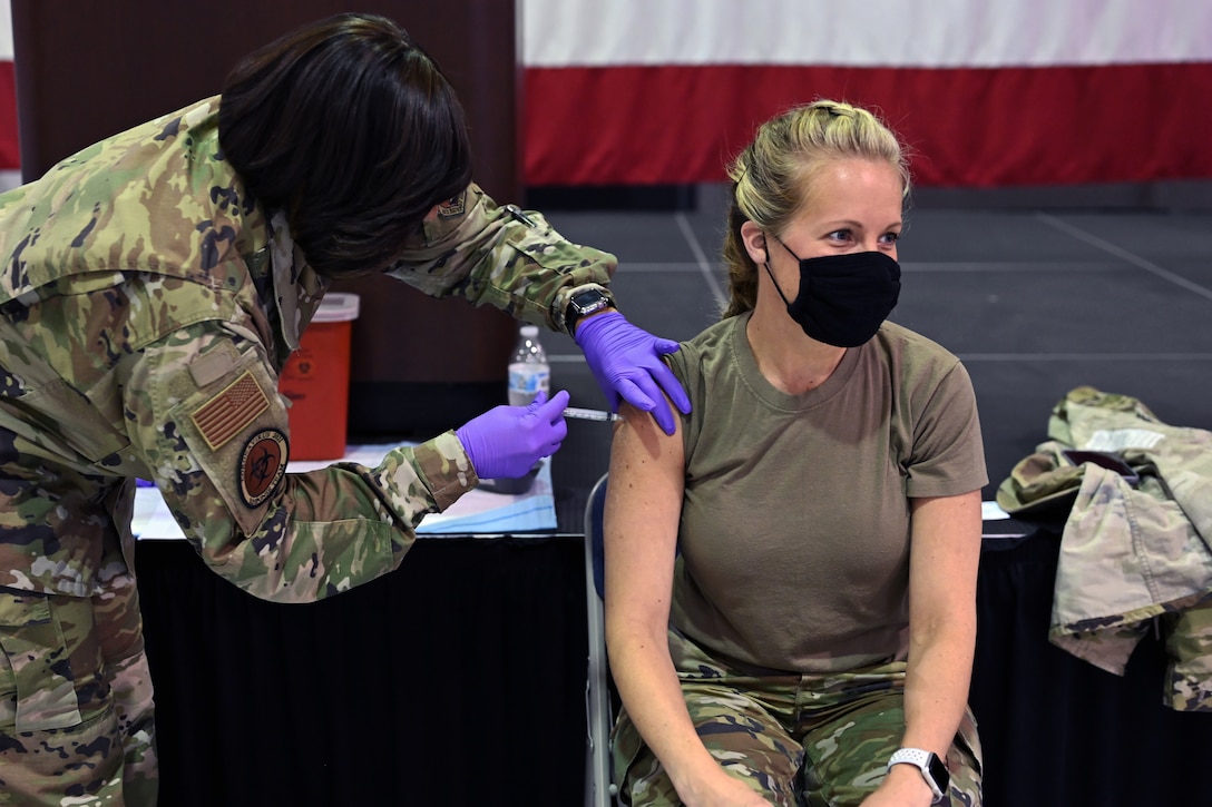 A soldier wearing a face mask and gloves leans down to give an injection into the arm of a woman who's seated and wearing a face mask.