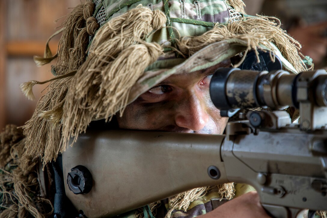 A soldier wearing a camouflage cover with mock foliage looks through the scope of a weapon.