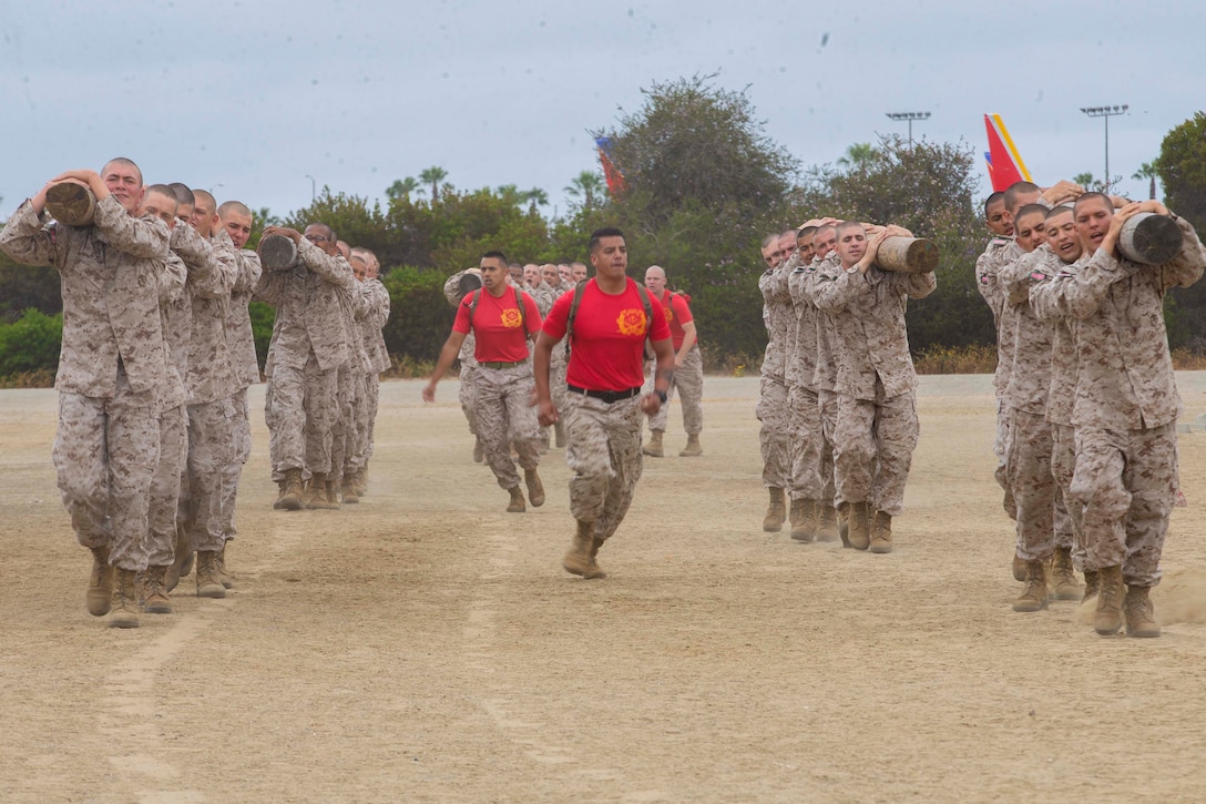 Marine Corps recruits carry logs as others walk/run beside them.