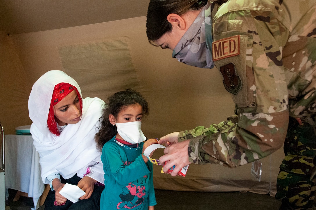 An airman hands an item to a child as a civilian adult watches.