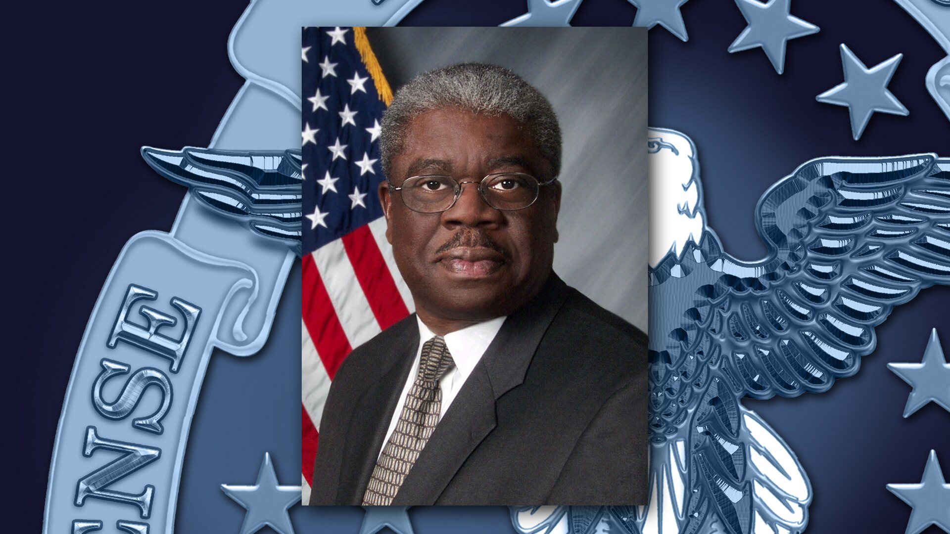 Black man in a dark suit sits in front of the US flag.