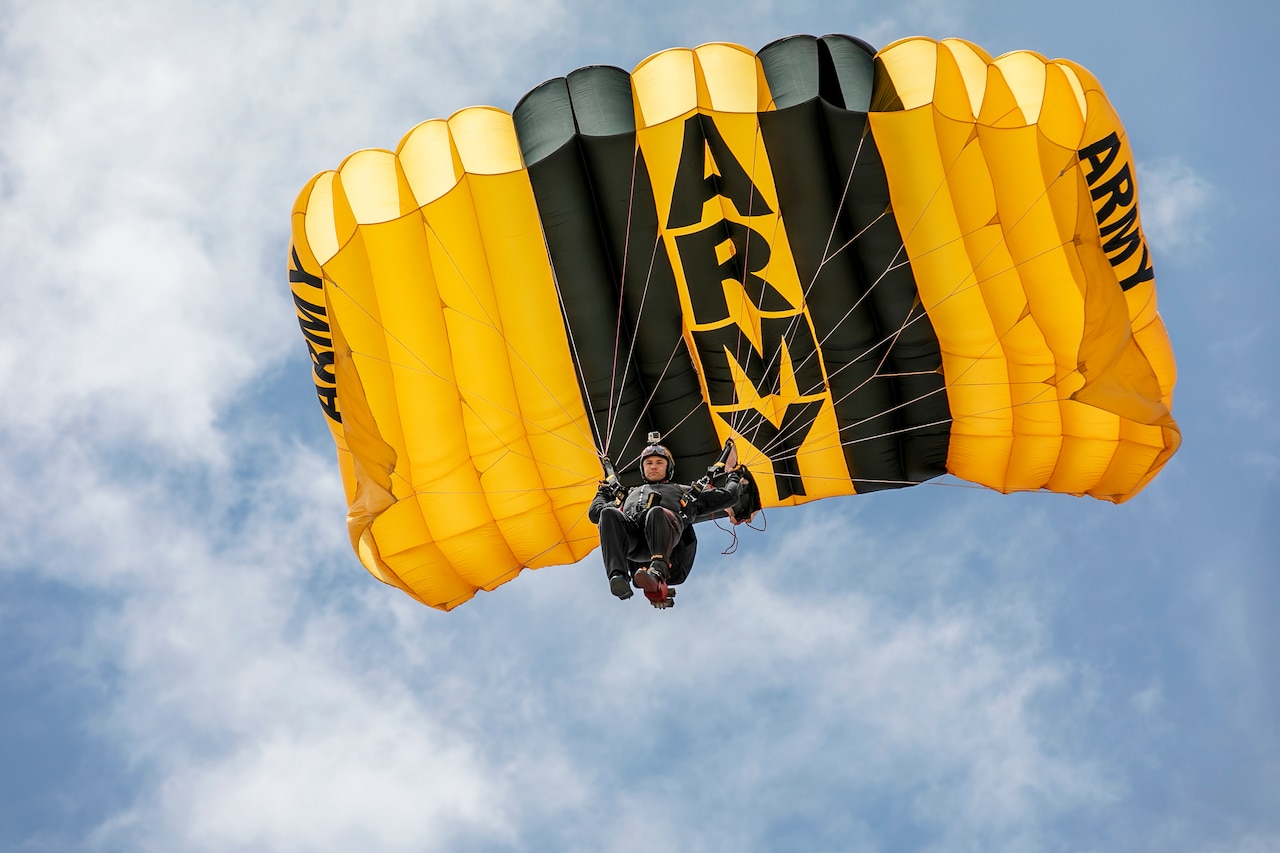 A soldier descends in blue sky with a gold and blue parachute with the word "ARMY" on it.
