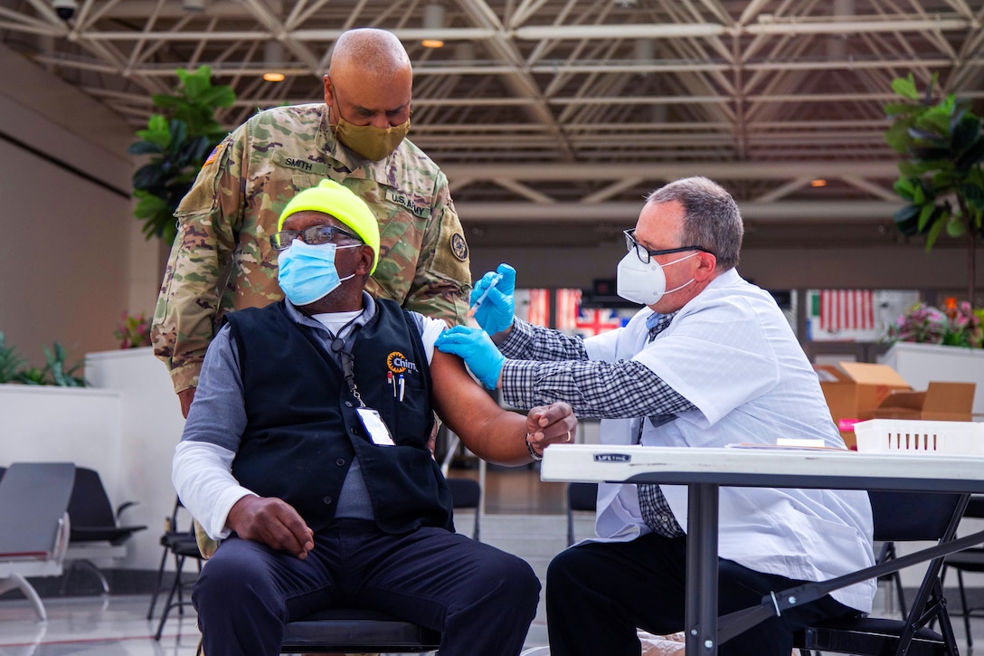 A soldier stands behind a man getting vaccinated.