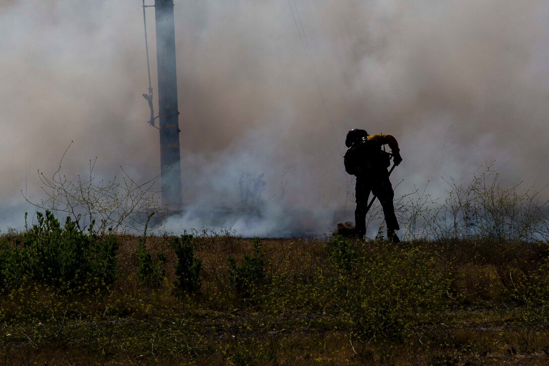 A person wearing firefighter gear digs near a grass fire as smoke surrounds.