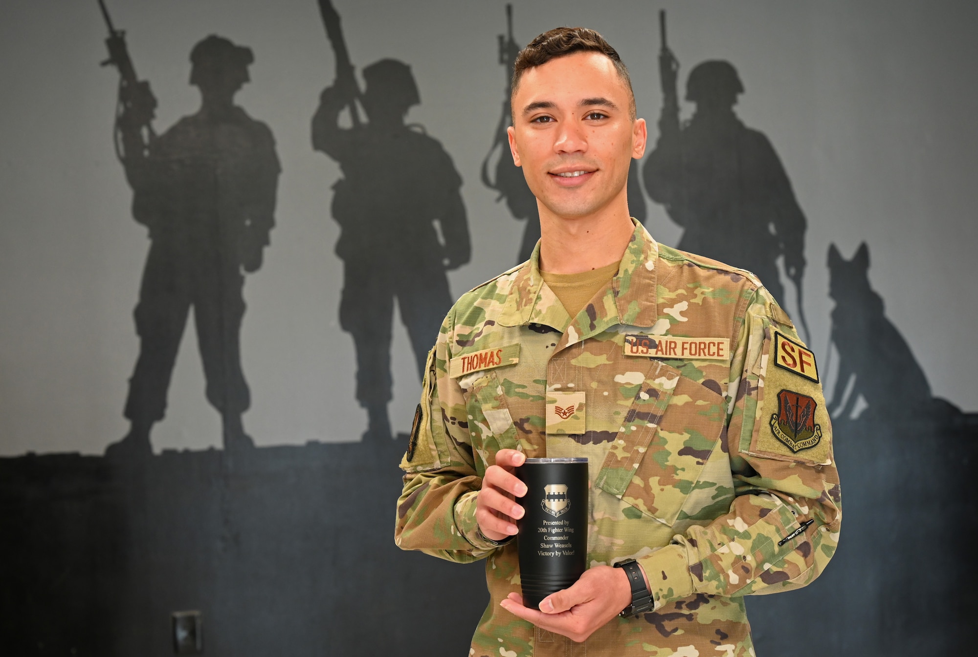 A photo of SSgt Thaddeus Thomas holds mug in front of mural.
