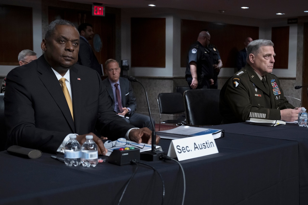 Secretary of Defense Lloyd J. Austin III and Army Gen. Mark A. Milley sit  in front of microphones at a table.