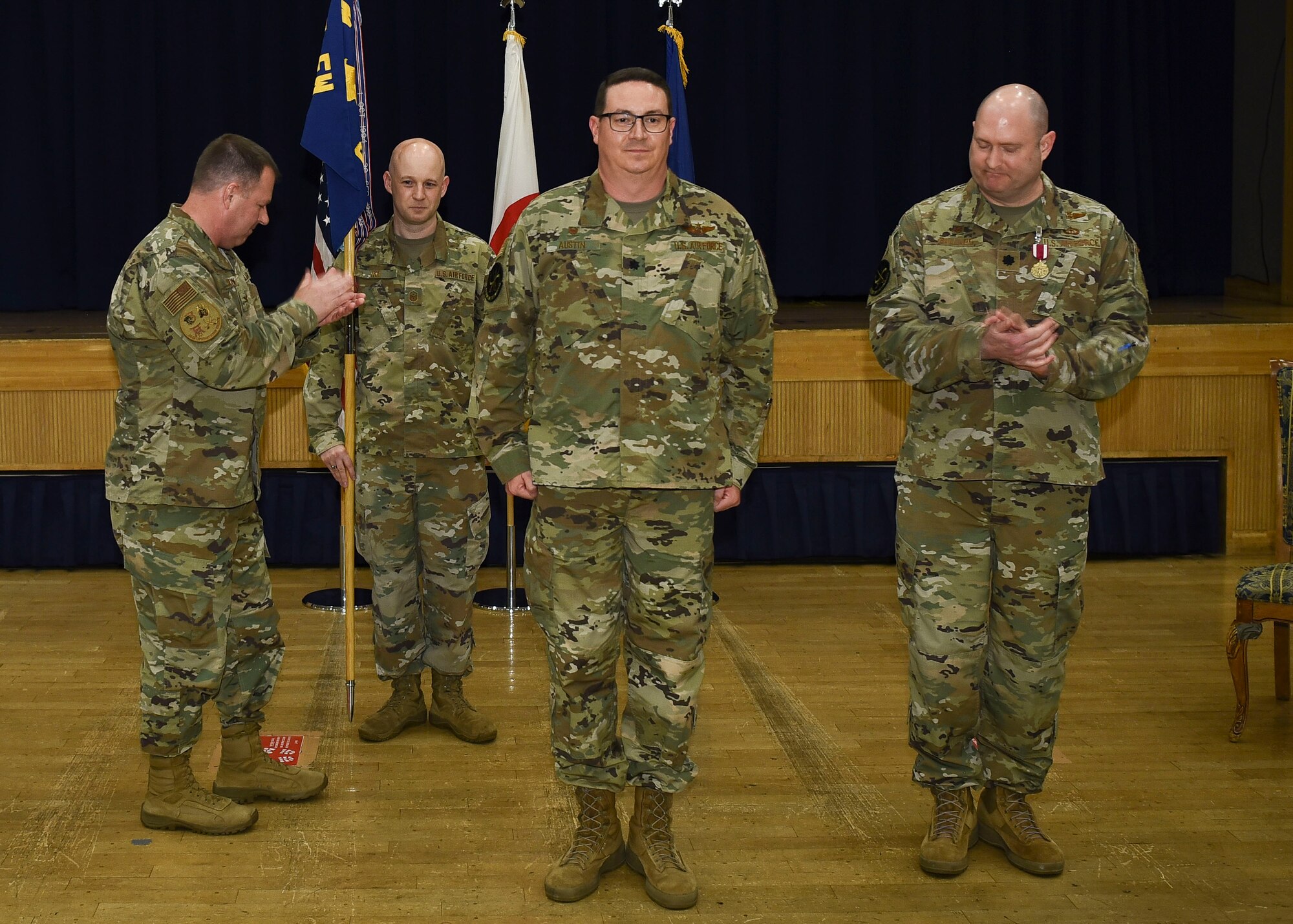 A military member holds a guidon while two other members clap for the center member standing in the position of attention.