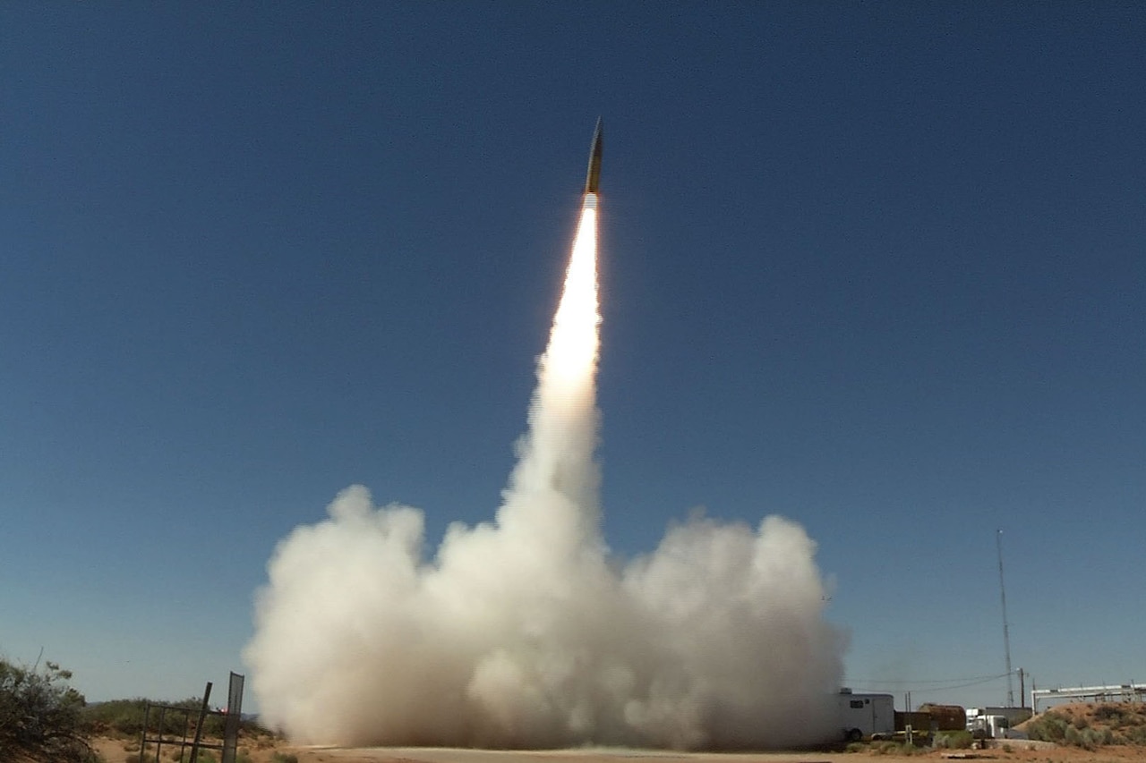 A missile launches skyward during a daytime test, leaving a trail of smoke and fire.