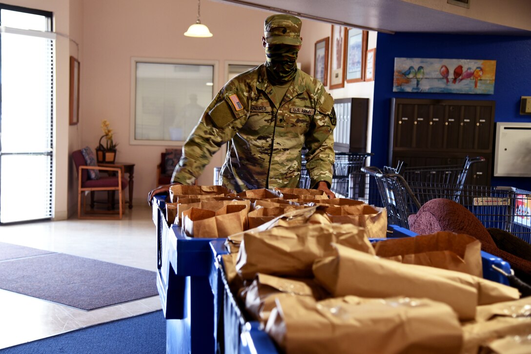 A service member wearing a face mask looks at two large bins filled with paper bags.