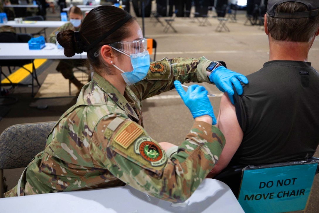 A female service member wearing a face mask and gloves gives an injection into the arm of a man seated next to her.
