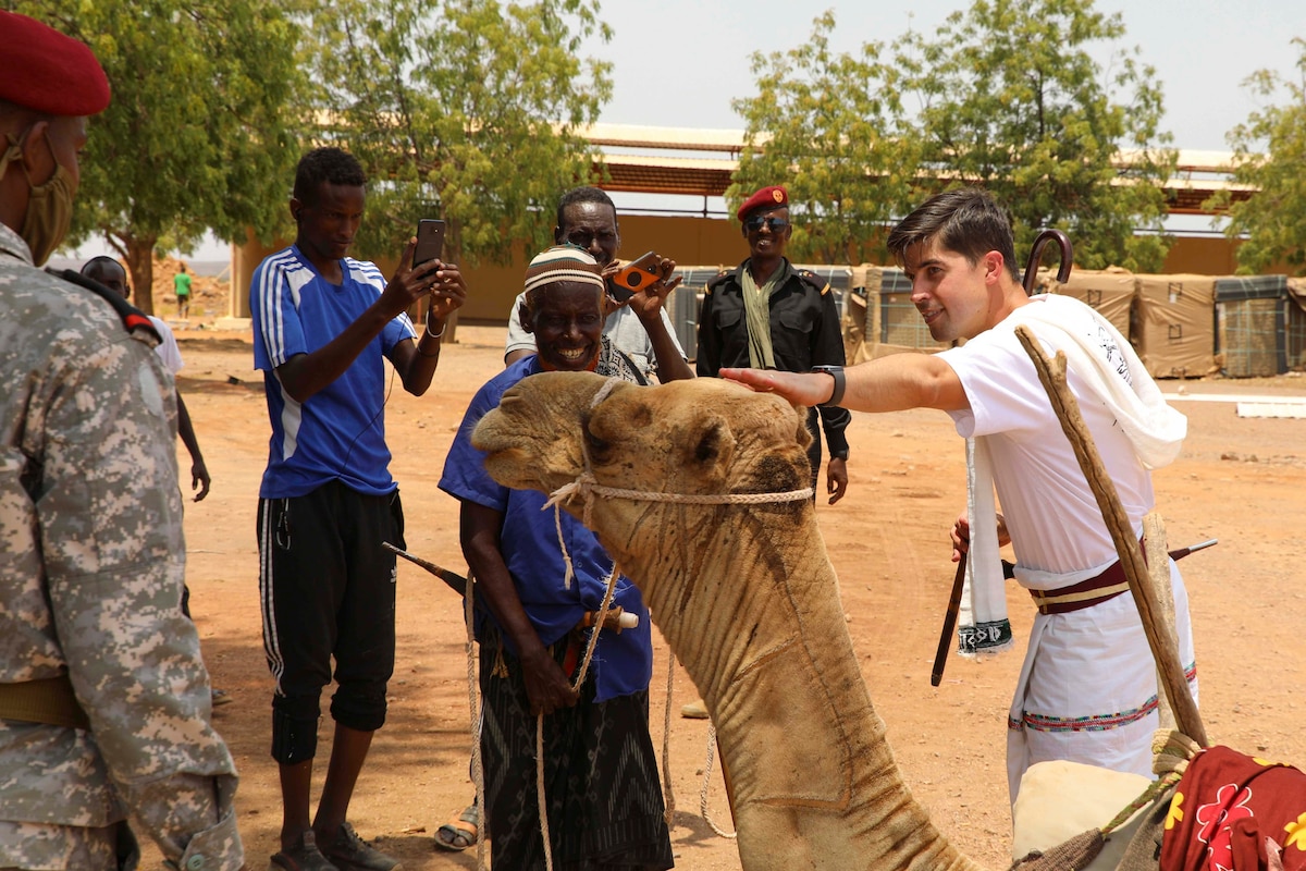 A soldier pets a camel as other peopole watch.