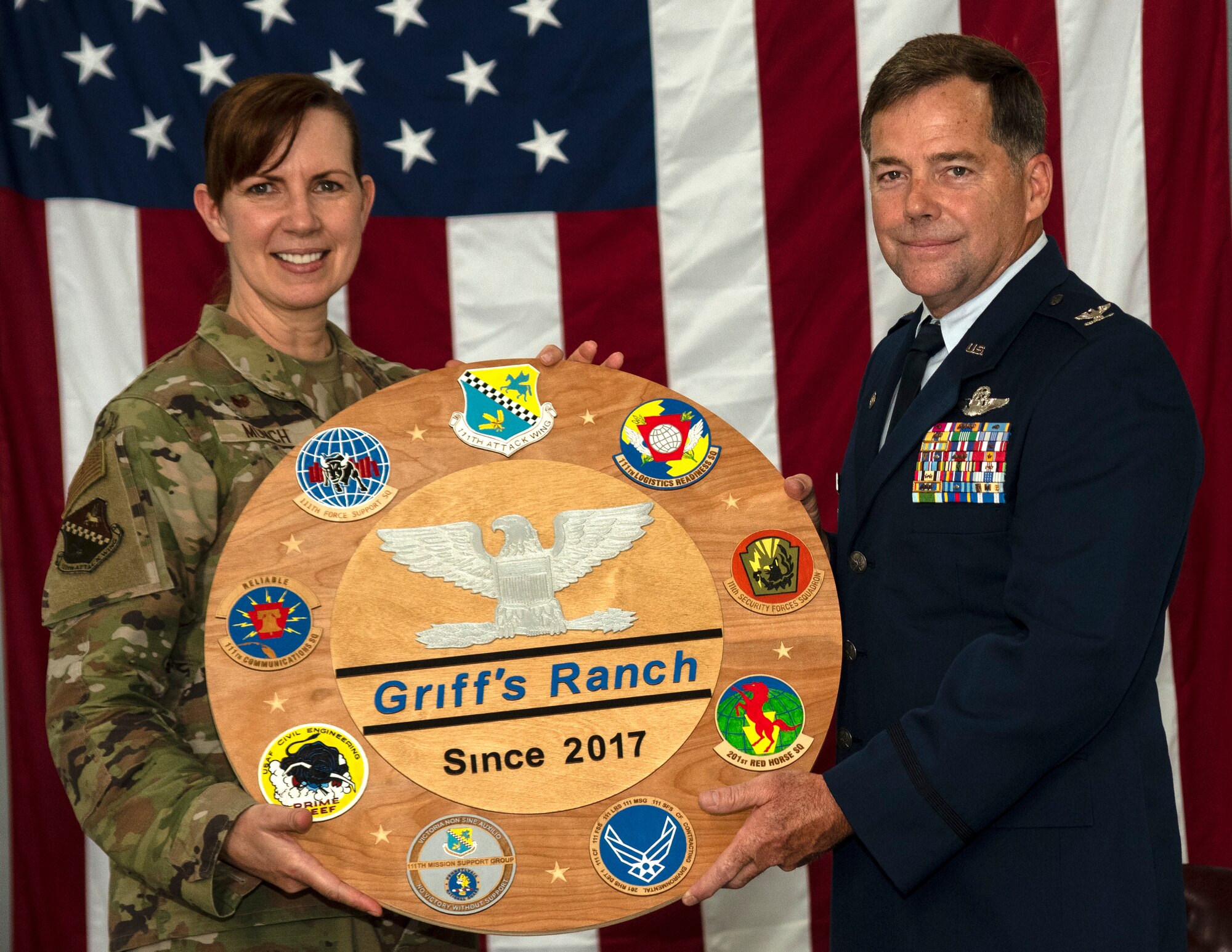 A woman wearing an Air Force camouflage uniform presents a plaque to a man wearing an Air Force Dress Blues.