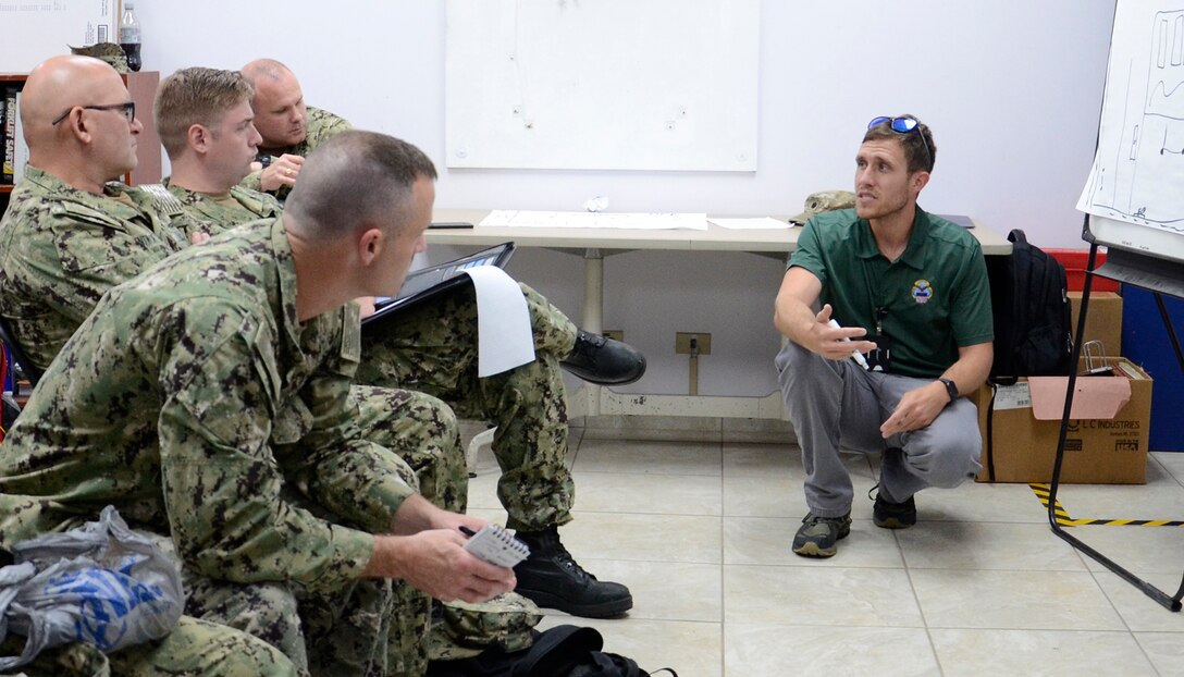 man kneels near poster board while talking to a group of military men seated.