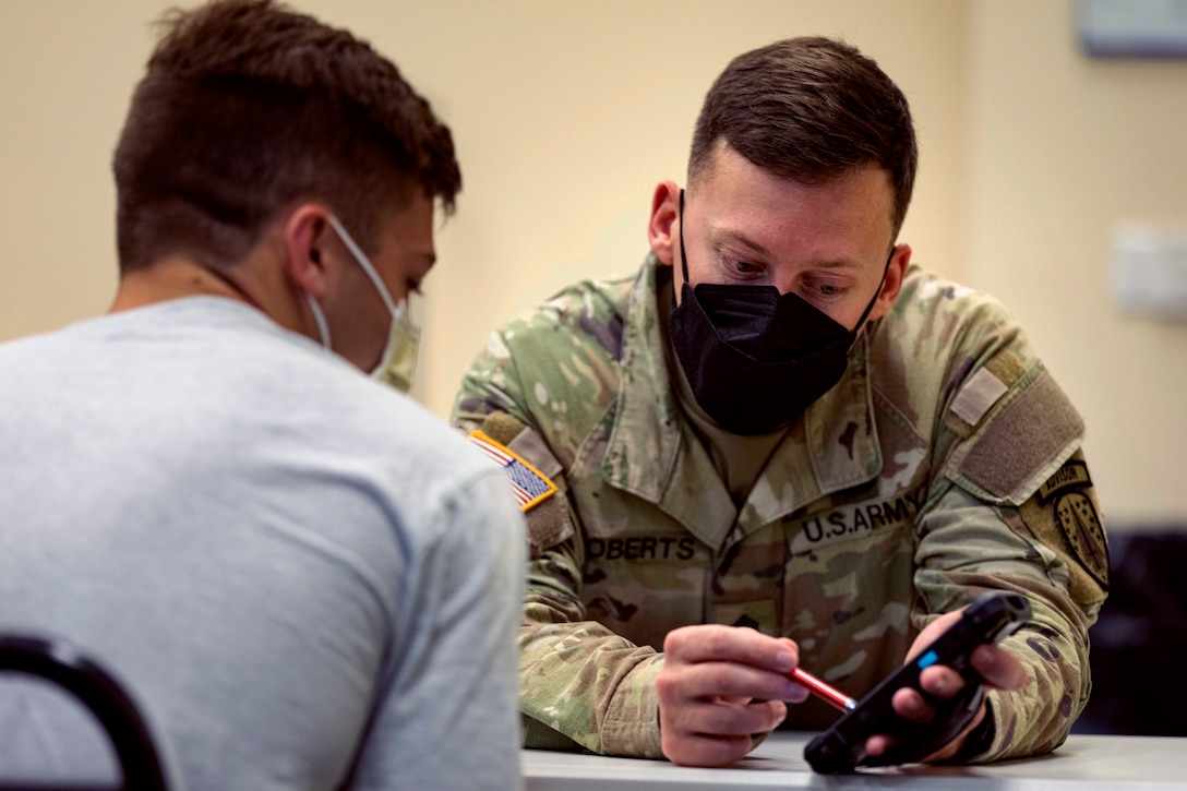 A soldier wearing a face mask sits at a table while screening a patient before administering a COVID-19 vaccine.