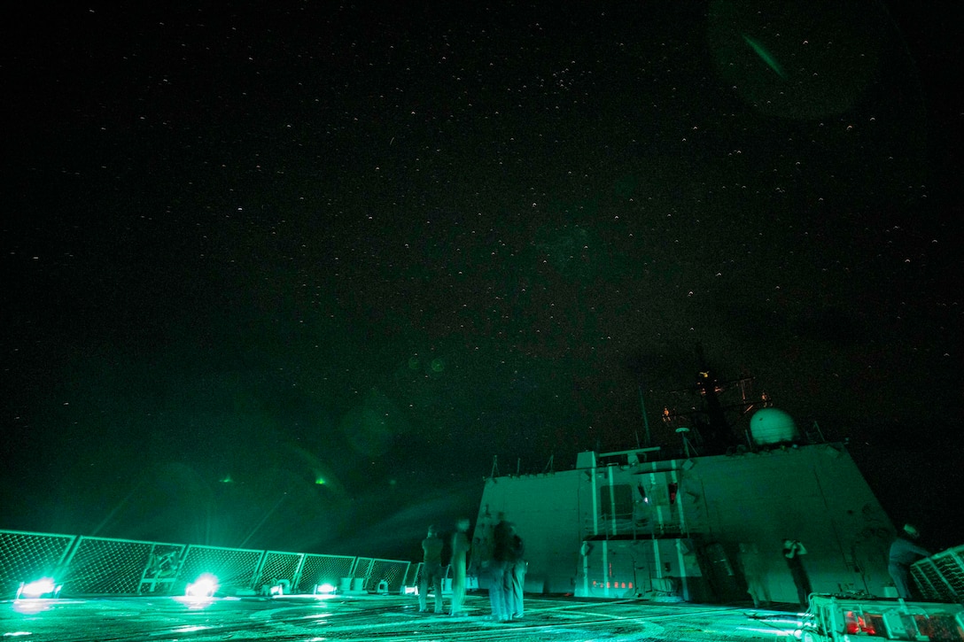 Sailors stand on a ship as it glows under starry night sky.