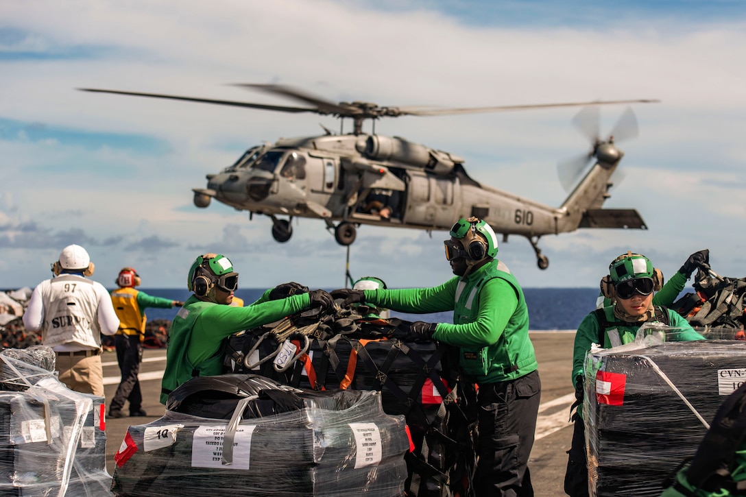 Sailors unstrap cargo on a ship at sea as a helicopter hovers above.