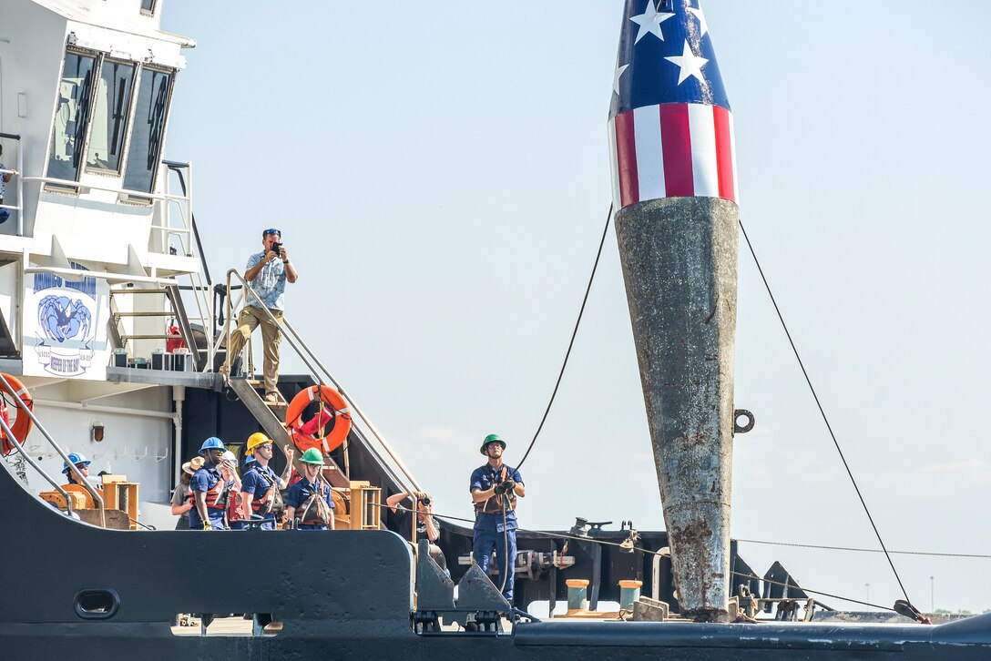 A Coast Guard crew member holds a line attached to a buoy painted with stars and stripes aboard a ship.