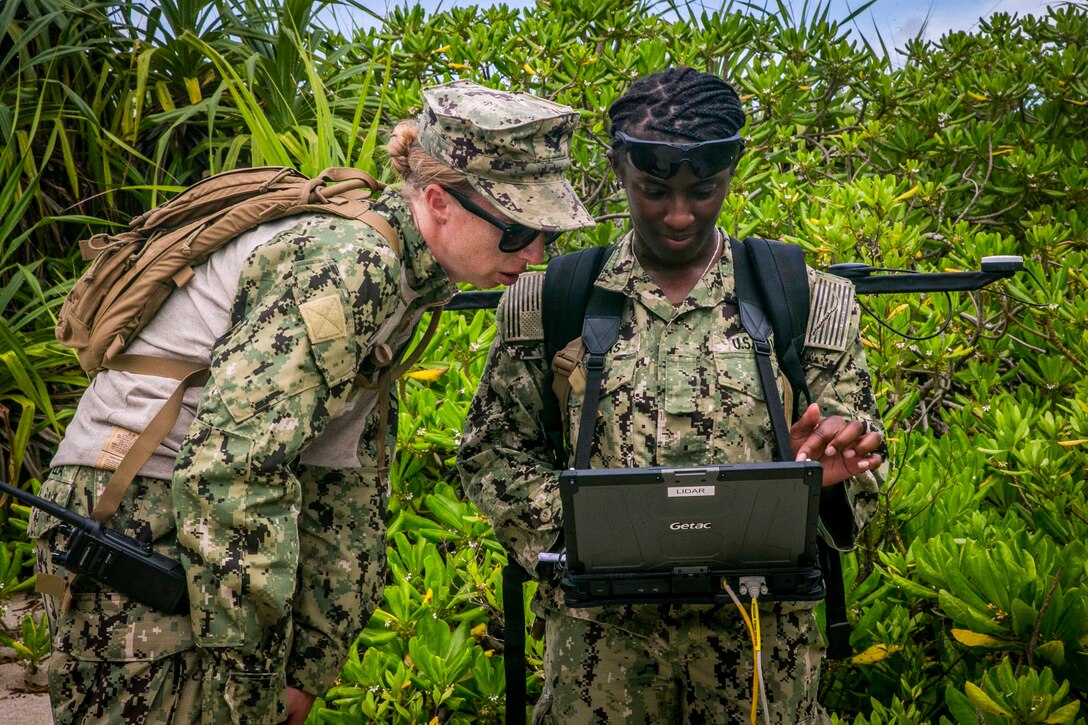 Two sailors look at a laptop screen in a jungle terrain.