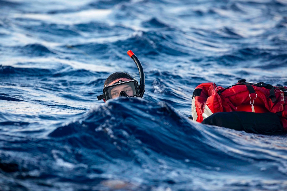 A sailor moves through waters above his shoulders next to a floating red bag.