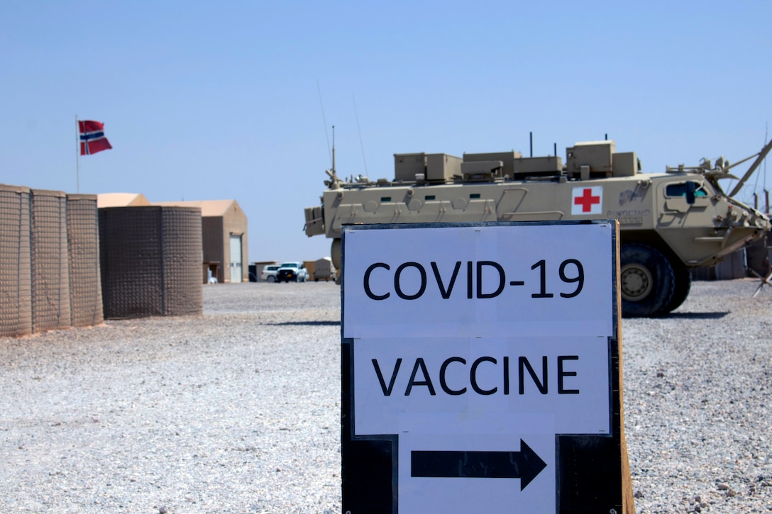 Against a dusty outdoor backdrop, a sign with the words "COVID-19 vaccine" has an arrow that points to the right.