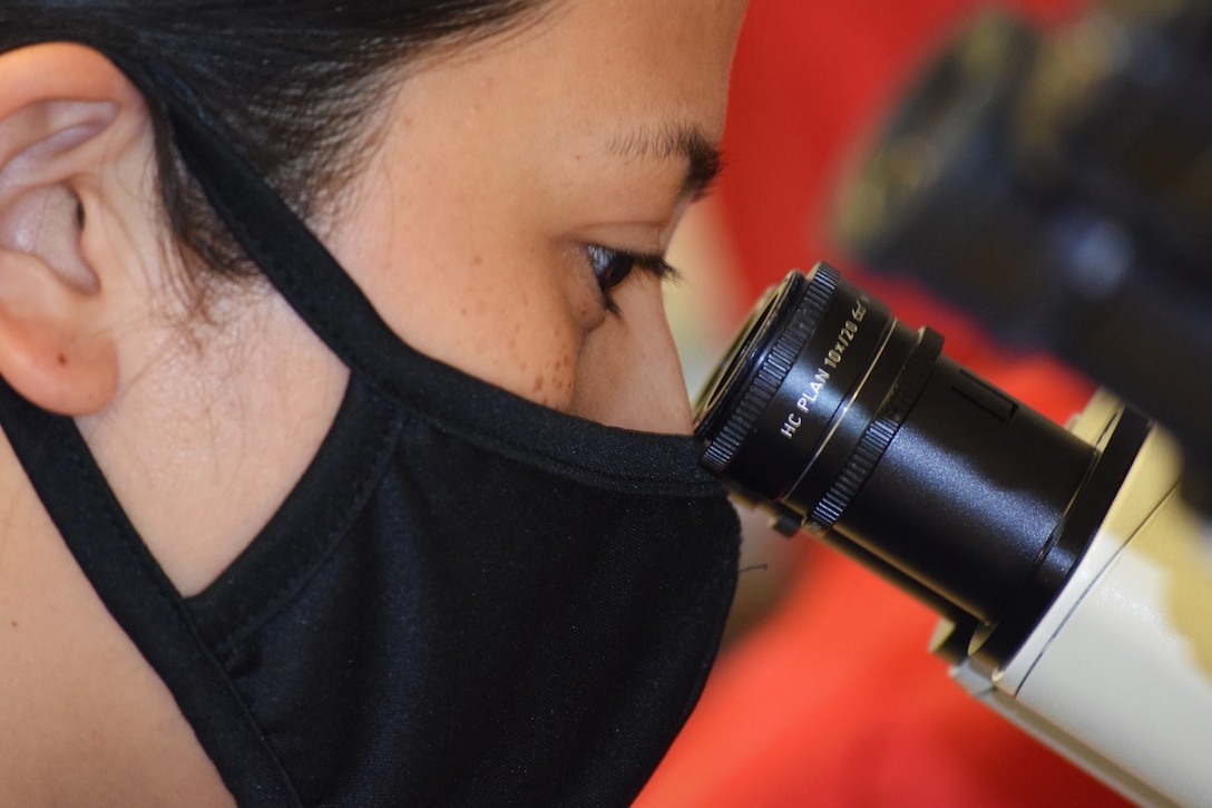 A woman wearing a face mask looks into a microscope.