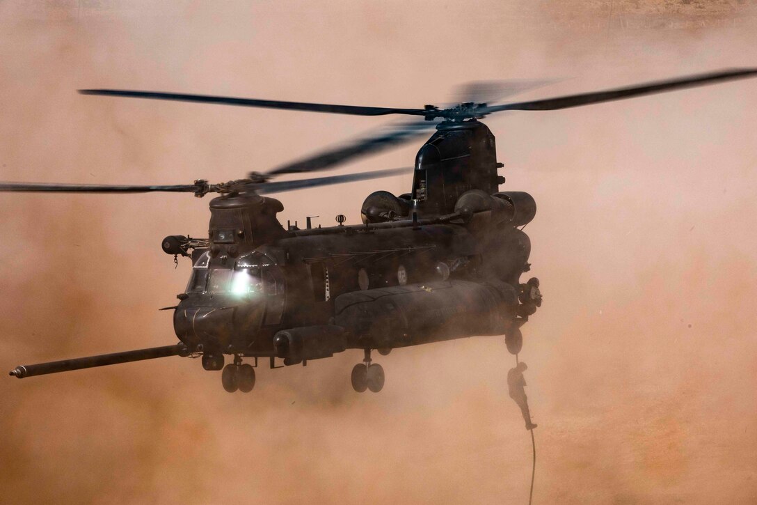 A soldier rappels from a helicopter as dust surrounds.