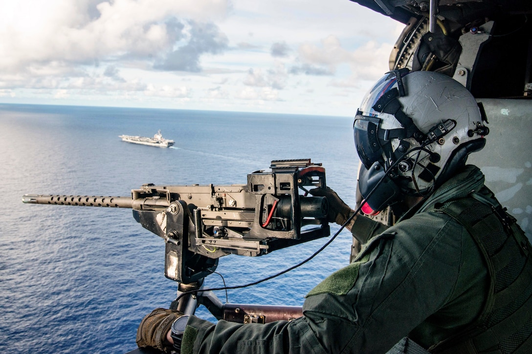 A sailor mans a machine gun aboard a helicopter while flying over water with a ship nearby.