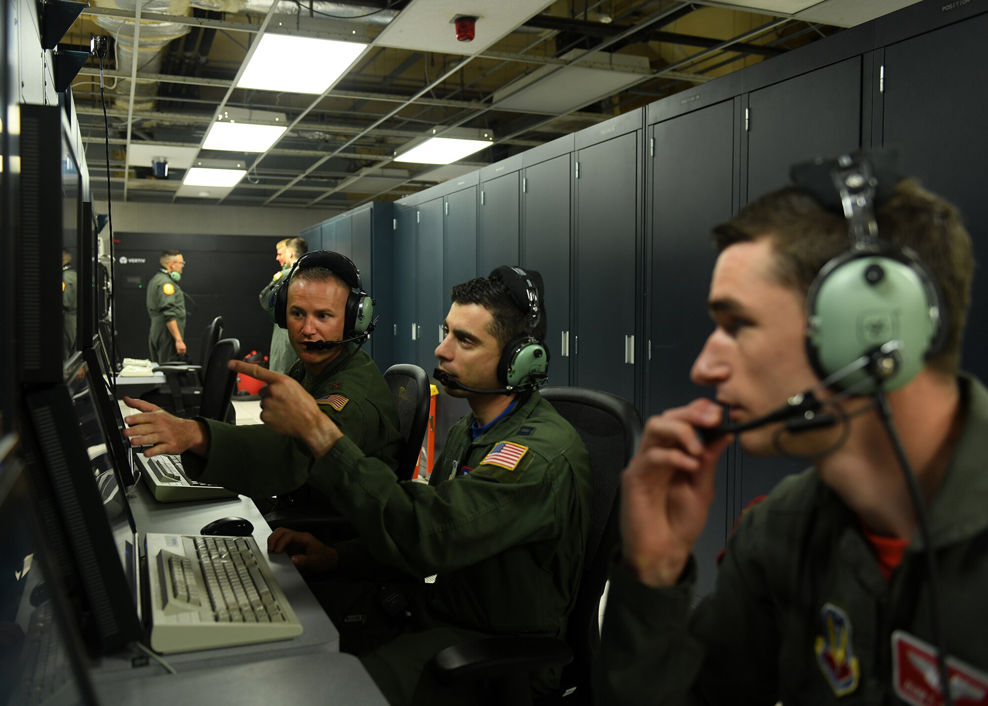 Three Airmen sit in front of a flight simulator terminal. Two men are pointing at computer screens.