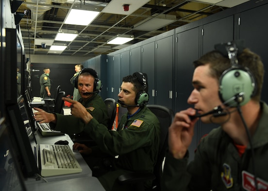 Three Airmen sit in front of a flight simulator terminal. Two men are pointing at computer screens.
