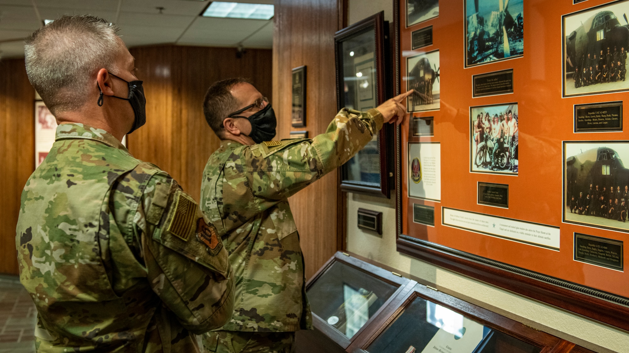 Two Airmen in uniform point at a photo on a wall.
