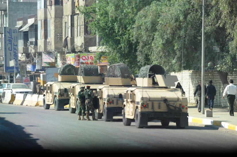 Humvees line up outside a tree-lined city street.