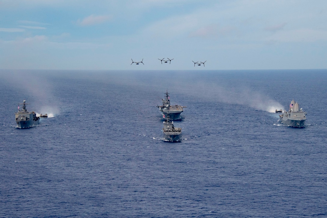 Four ships and two landing craft sail in formation as three aircraft fly above.
