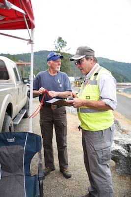 Two men, one with clipboard, one with rope tied around a pole.