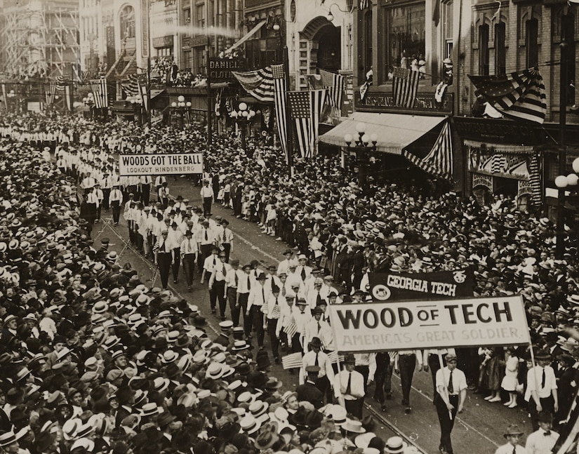 A band marches through a street packed with spectators. A sign at the front says, "Wood of Tech: America’s great soldier."