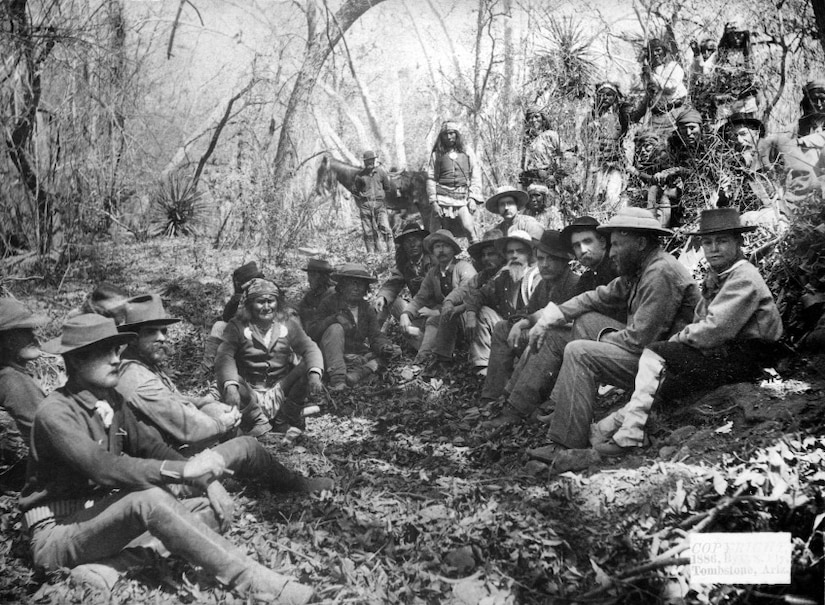 Several men sit on the ground in a wooded area.