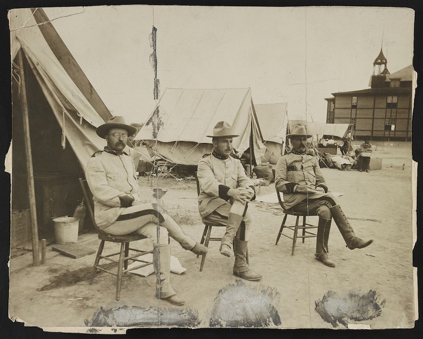 Three men sit on wooden chairs with tents behind them.