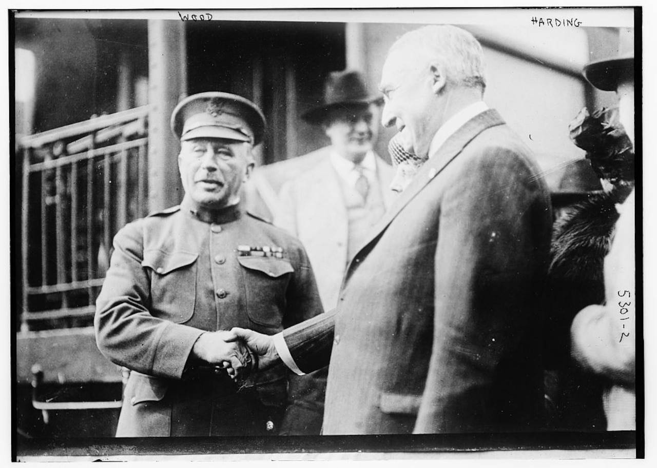A soldier in dress uniform shakes the hand of a man in a suit.